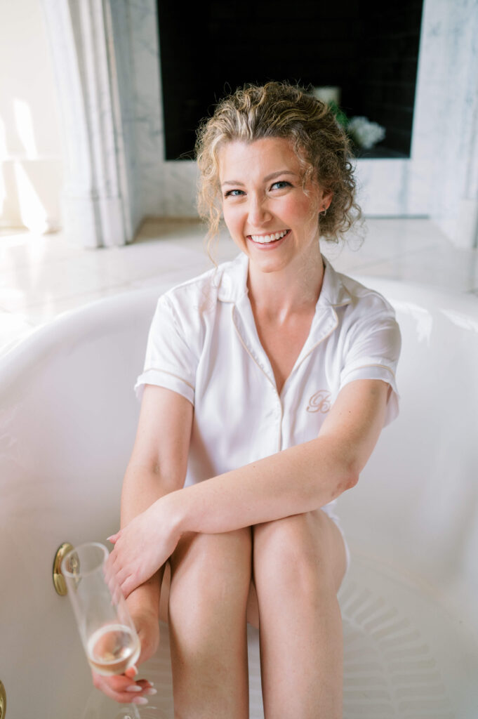 a lovely bride sits in a grand tub with a glass of champagne on her wedding day