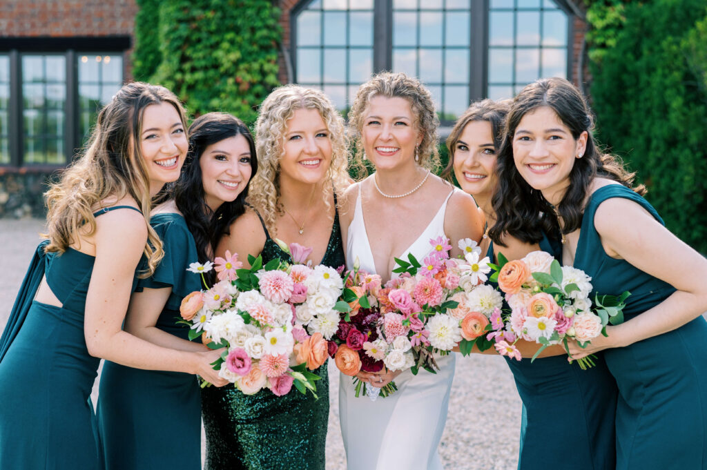 Beautiful bridesmaids in shades of green hold pink and orange flowers while standing next to their bride at Dover Hall Estate