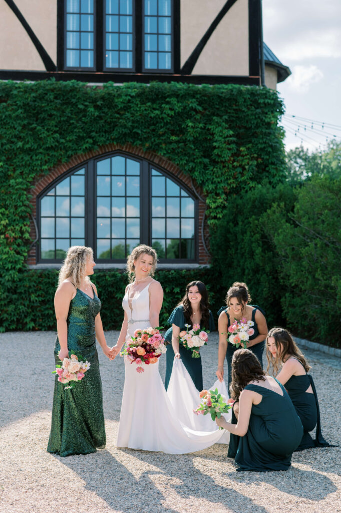 Bridesmaids in green dresses adjust the train of blonde bride with curly hair while standing in front of a vine covered brick wall