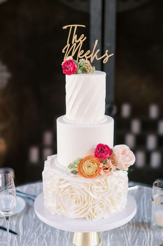 A beautiful three tiered white cake adorned with pink and orange flowers sits atop a table at a wedding