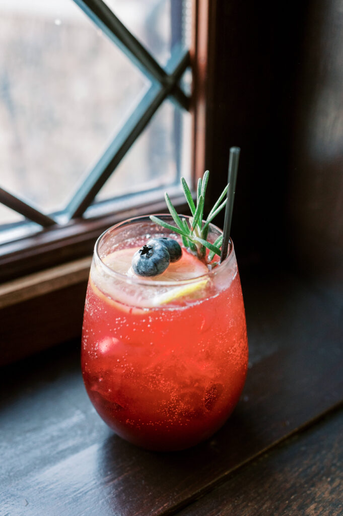 A beautiful cocktail in shades of red and topped with blueberries and rosemary is passed around during cocktail hour at a wedding