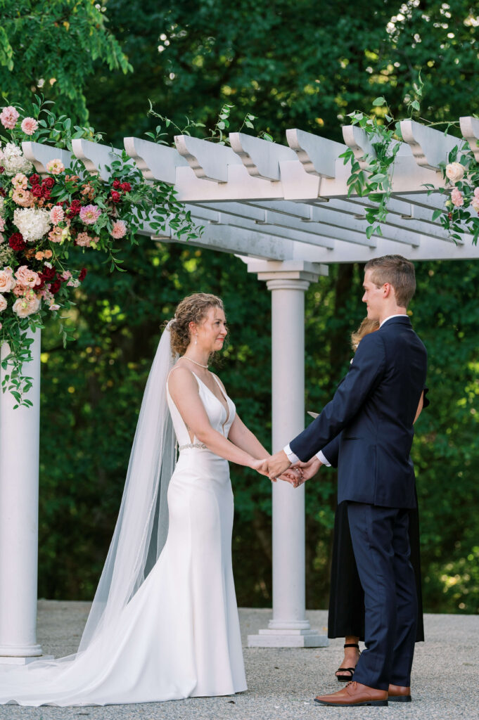 A bride wears a slim white satin dress and holds the hands of her groom who is wearing a navy suit while they stand at the alter during their wedding