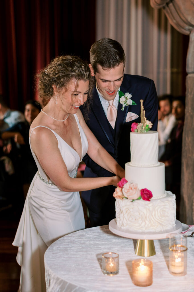 The bride and groom smile and laugh while cutting the three tiered white cake at their wedding