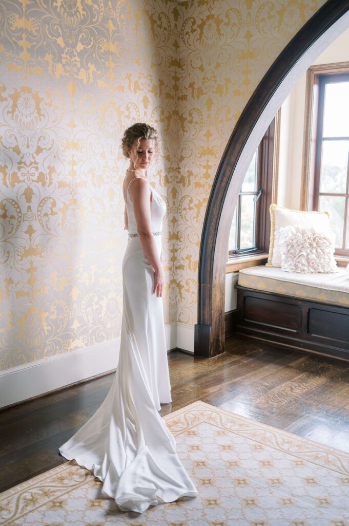 A beautiful bride in a slim white satin wedding dress looks over and down her shoulder while getting dressed for her wedding at Dover Hall Estate