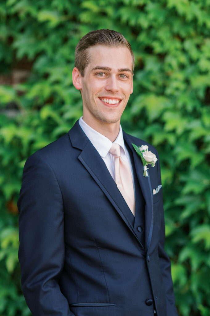 Handsome groom in a navy suit and pink tie smiles at the camera during his wedding