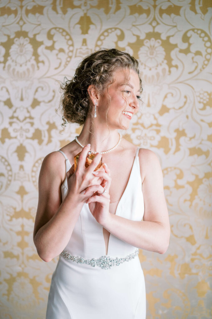 A blonde bride smiles wide while putting on perfume before her wedding